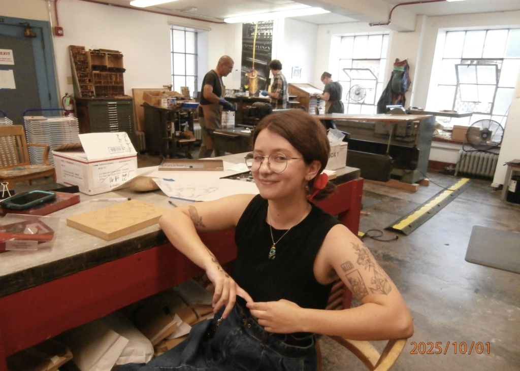 Chloë, wearing a black tank top and jeans, sits in front of a bright red desk that is covered in assorted printing materials